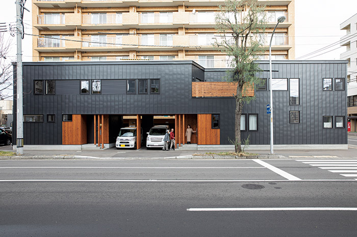 片側二車線の通り沿いに建つKさん宅。黒のガルバリウム鋼板に木板張りを組み合わせた横長の形状は存在感あり。二世帯のうちKさんご夫妻と娘さんが暮らすのは左側の部分
